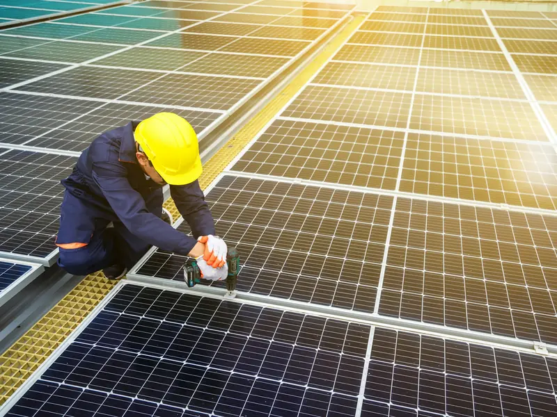 Installer in a hard hat and gloves fixing solar panels to their mounting rails on a roof